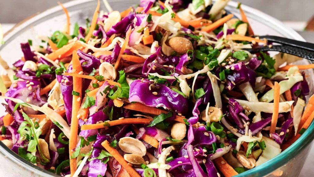 A bowl of colorful cabbage salad with shredded red and green cabbage, carrots, green onions, peanuts, sesame seeds, and herbs.
