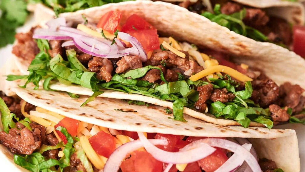 Close-up of three tacos filled with ground beef, shredded lettuce, diced tomatoes, sliced red onions, and shredded cheese in flour tortillas.