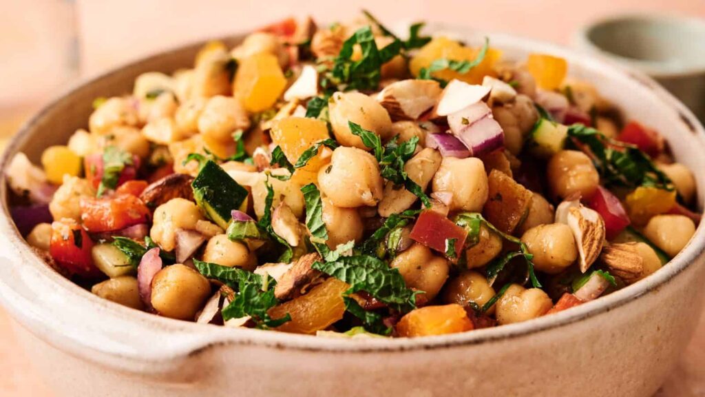 A bowl of chickpea salad with chopped vegetables, herbs, nuts, and dried fruit, served in a ceramic dish.