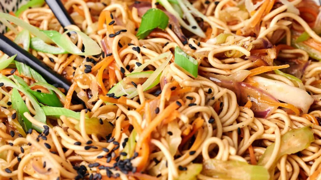 Close-up of stir-fried noodles with sliced green onions, shredded carrots, vegetables, and black sesame seeds.