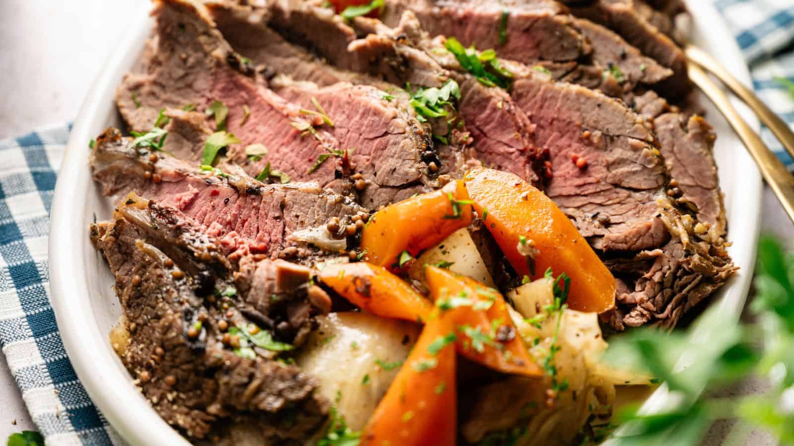 Sliced roast beef served on a plate with cooked carrots, potatoes, and herbs, placed on a checkered cloth.