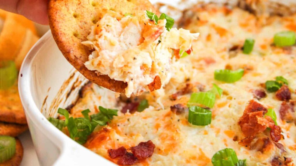 A cracker dipped into a baked cheesy dip topped with green onions and bacon bits.