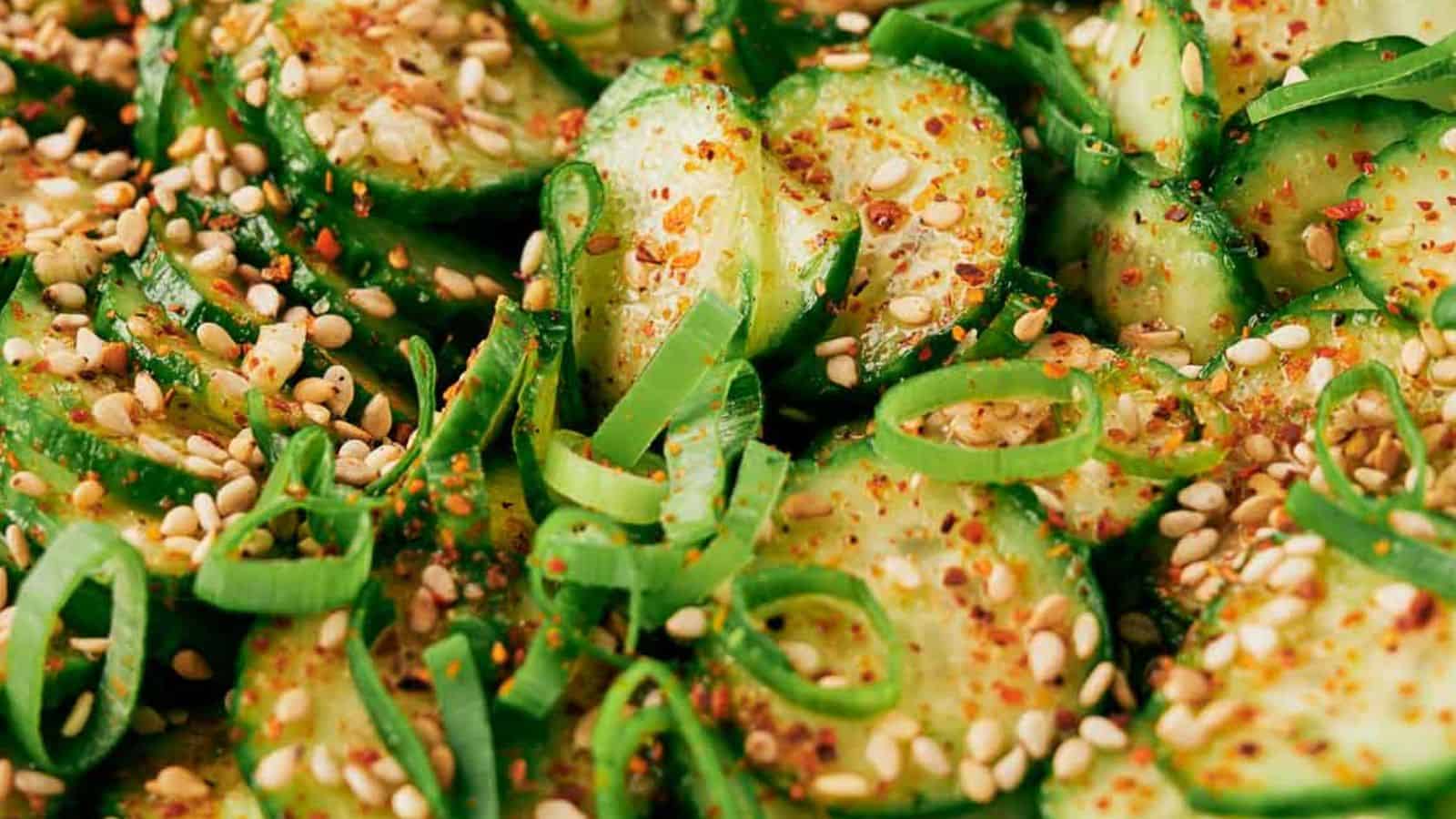 Close-up of sliced cucumbers garnished with chopped scallions, sesame seeds, and red chili flakes.