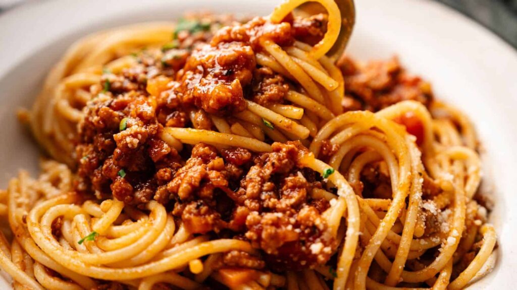 A close-up of spaghetti topped with meat sauce and garnished with grated cheese and herbs, served on a white plate.