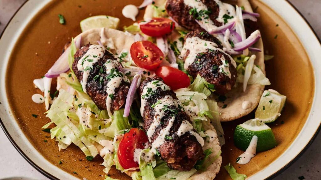 Three grilled kebabs on flatbread with shredded lettuce, sliced red onion, cherry tomatoes, lime wedges, and a drizzle of white sauce on a brown plate.