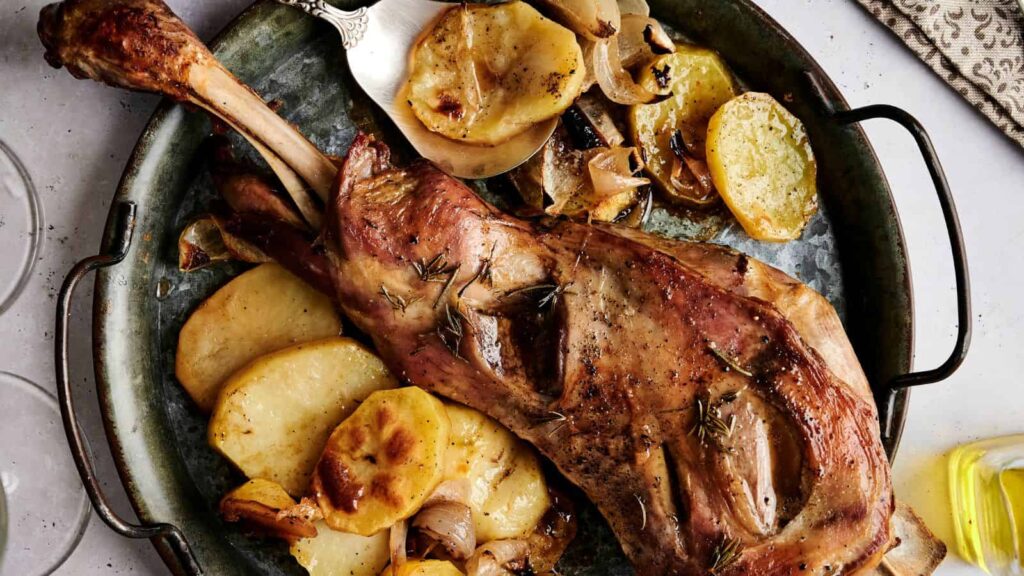 A roasted leg of lamb on a metal tray, surrounded by sliced potatoes and onions, garnished with rosemary sprigs.