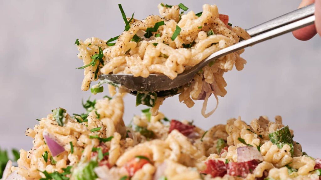 A spoon scoops up a serving of macaroni salad with vegetables and herbs, highlighted by soft lighting.