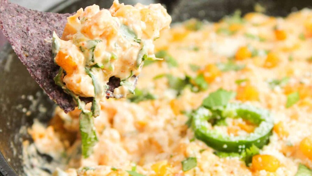 A close-up of a spoonful of cheesy dip with chopped green herbs and peppers, held above a bowl of the same mixture.