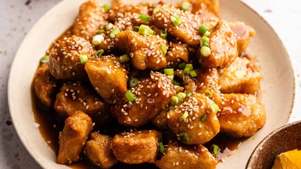 A plate of orange chicken pieces covered in sauce, garnished with sesame seeds and chopped green onions.