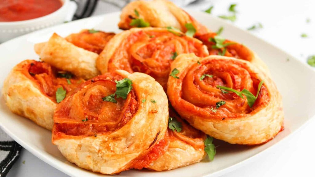 A plate of puff pastry pizza rolls garnished with cilantro, placed on a white surface. A bowl of tomato sauce is partially visible in the background.