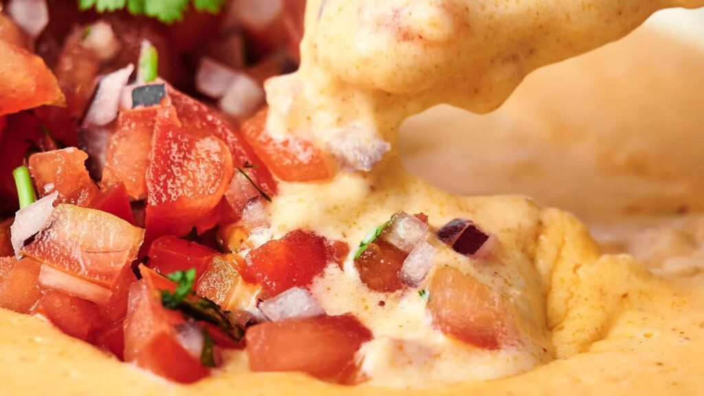 Close-up of creamy cheese dip being scooped with a mix of chopped tomatoes, onions, and herbs on top.
