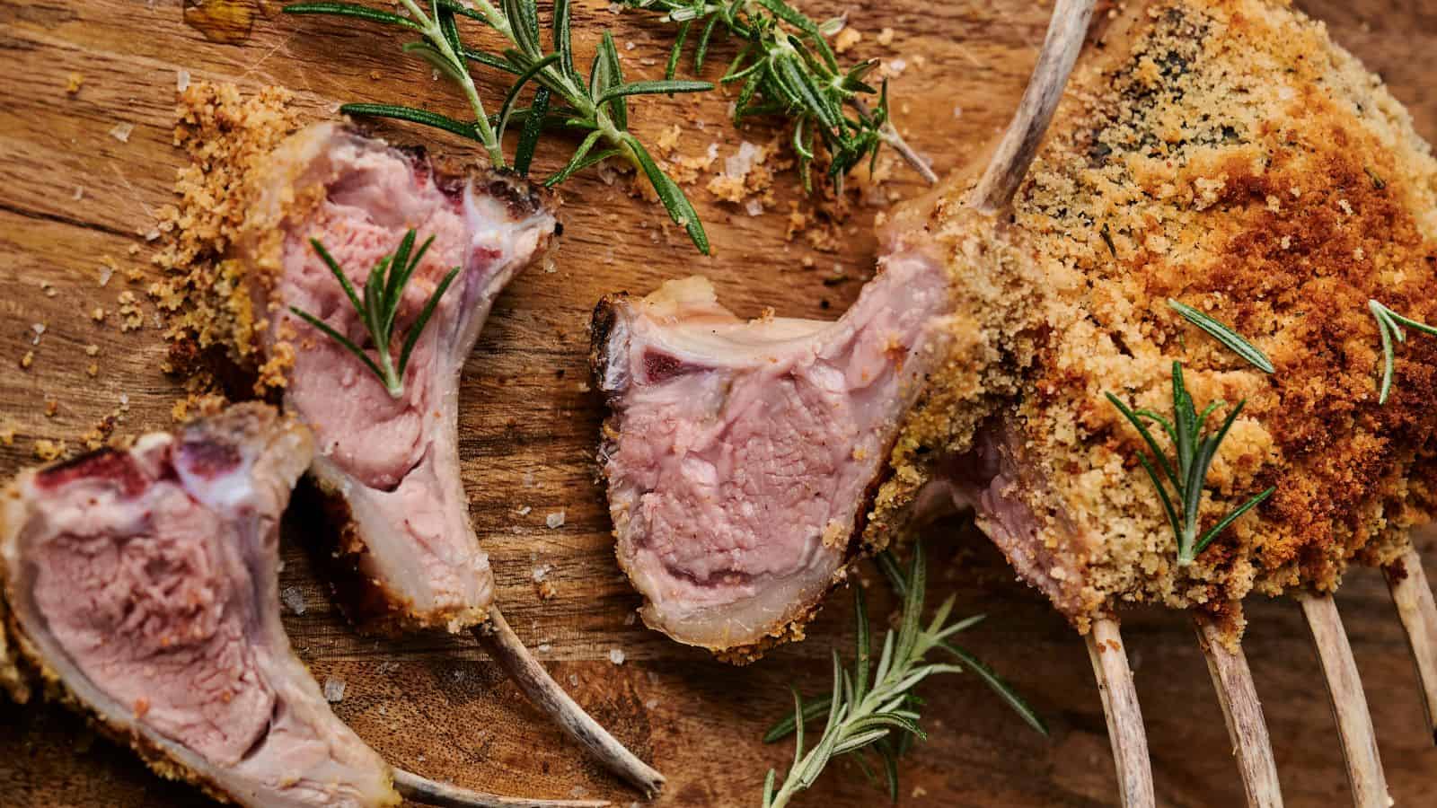 Rack of lamb with a breadcrumb crust is garnished with rosemary on a wooden board. Sliced pieces reveal a pink, cooked interior.