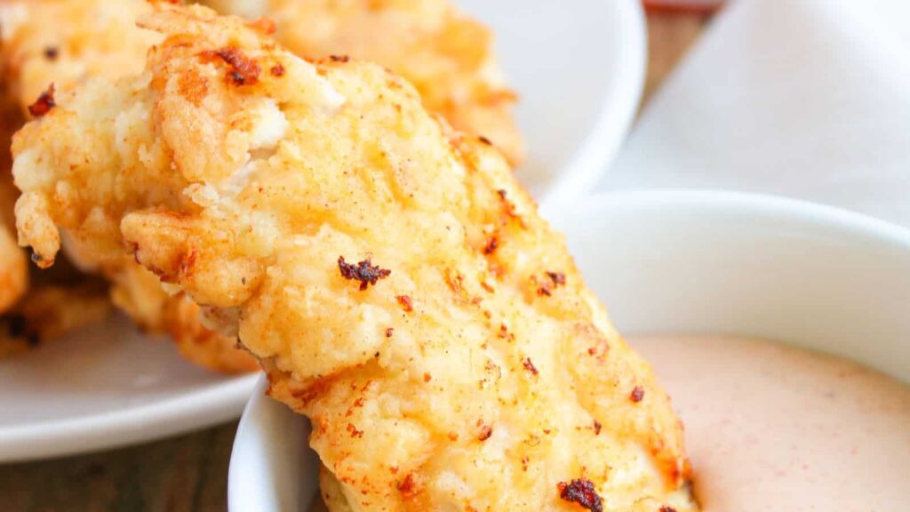 Close-up of a golden-brown, crispy chicken strip being dipped into a bowl of sauce.
