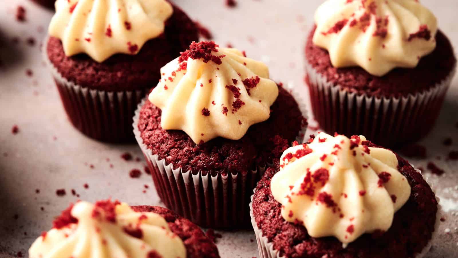 Several red velvet cupcakes with swirls of cream cheese frosting and sprinkled crumbs on top, arranged on a light-colored surface.