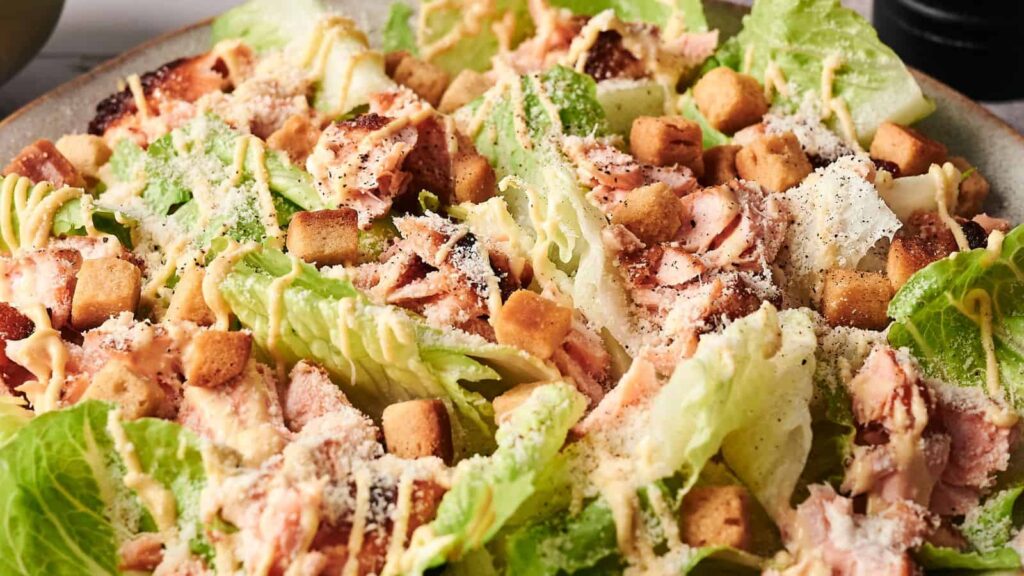 Close-up of a Caesar salad with lettuce, grilled chicken, croutons, grated cheese, and dressing on a plate.
