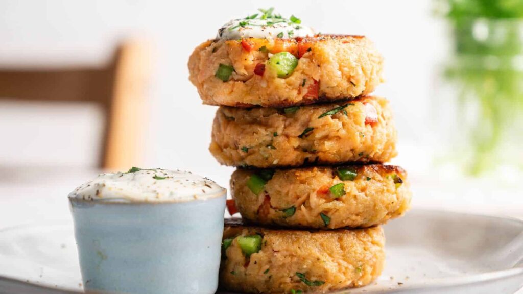 A stack of four salmon patties is plated next to a small cup of creamy dipping sauce, garnished with herbs.
