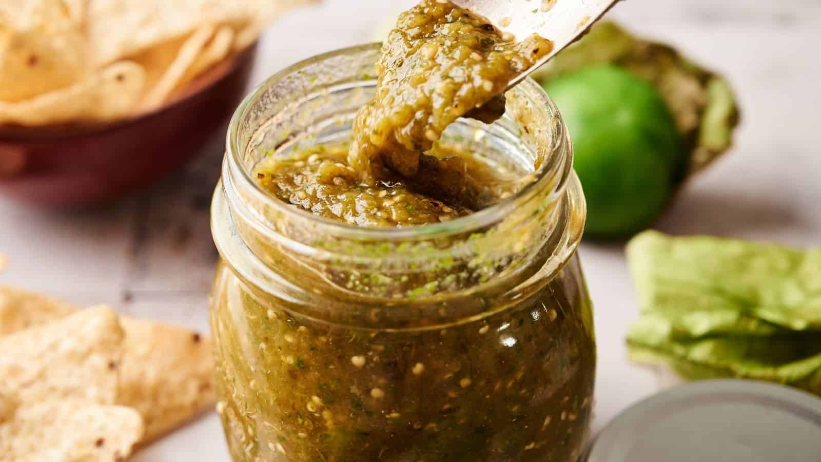 A jar of green salsa with a spoon lifting some out; tortilla chips and a tomatillo are visible in the background.