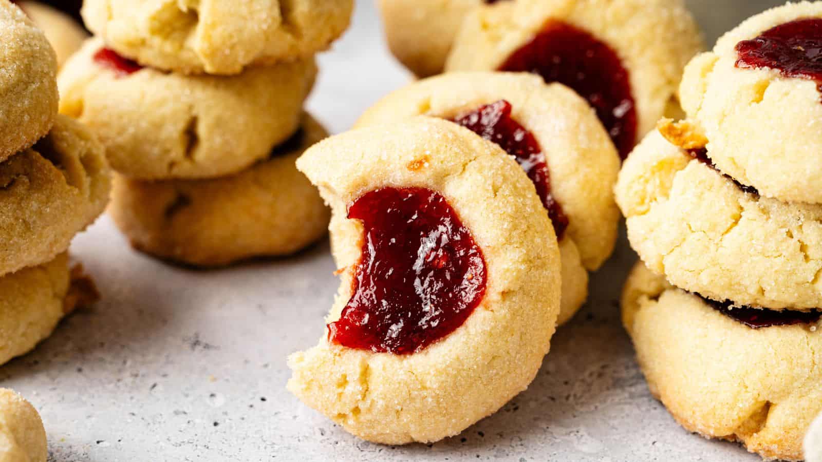 Close-up of several stacks of thumbprint cookies filled with red jam, with one cookie in the foreground showing a bite taken out.