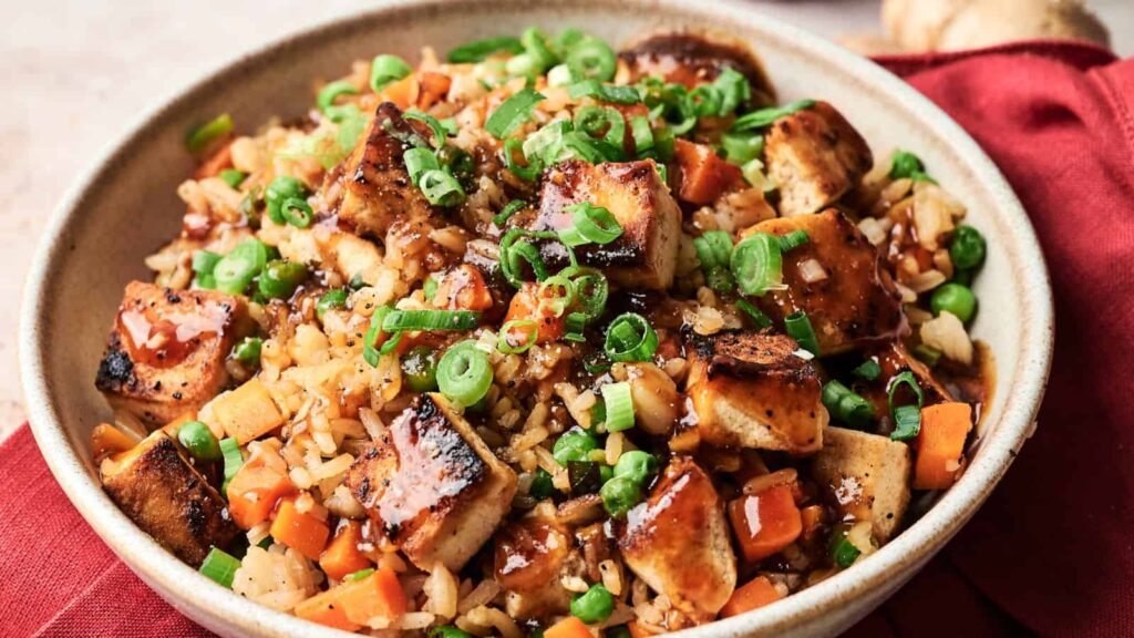 A bowl of fried rice with grilled tofu cubes, diced carrots, green peas, chopped green onions, and a drizzle of sauce.