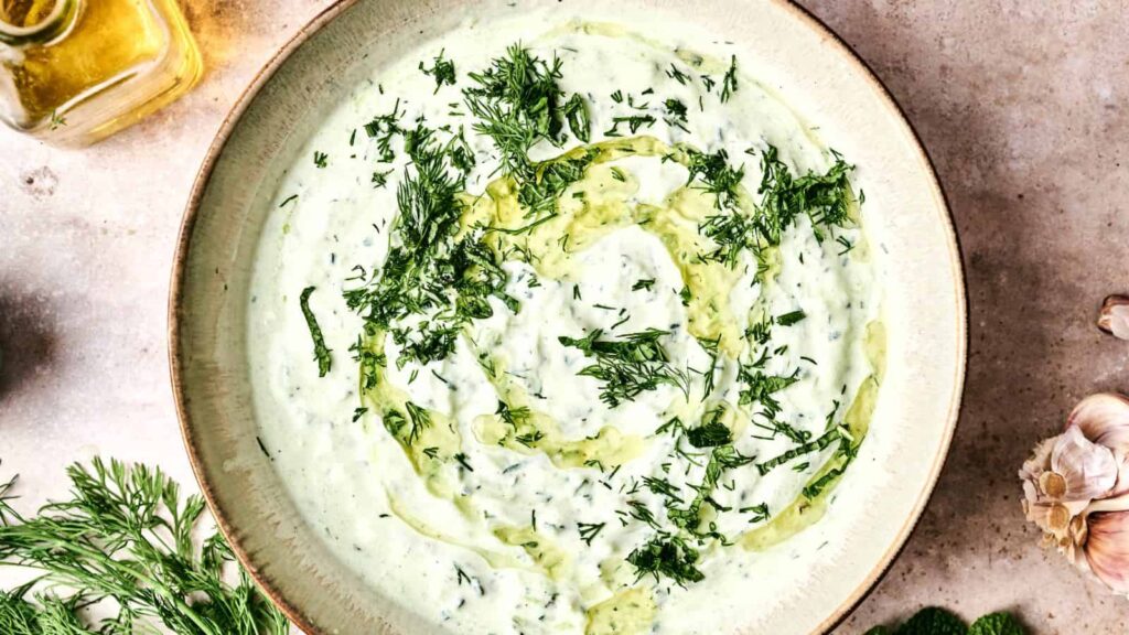 A bowl of tzatziki sauce garnished with fresh dill and a swirl of olive oil, placed on a stone surface with garlic and dill nearby.