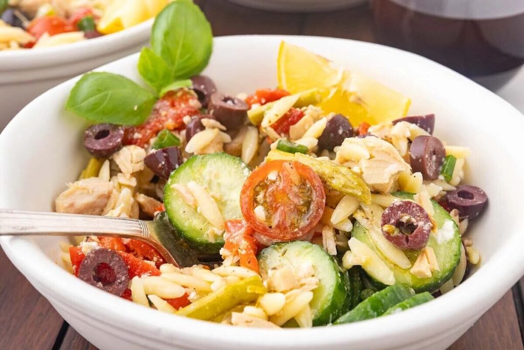 A bowl of Mediterranean tuna orzo salad with sliced cucumbers, cherry tomatoes, olives, herbs, and a lemon wedge. A fork is placed inside the bowl.