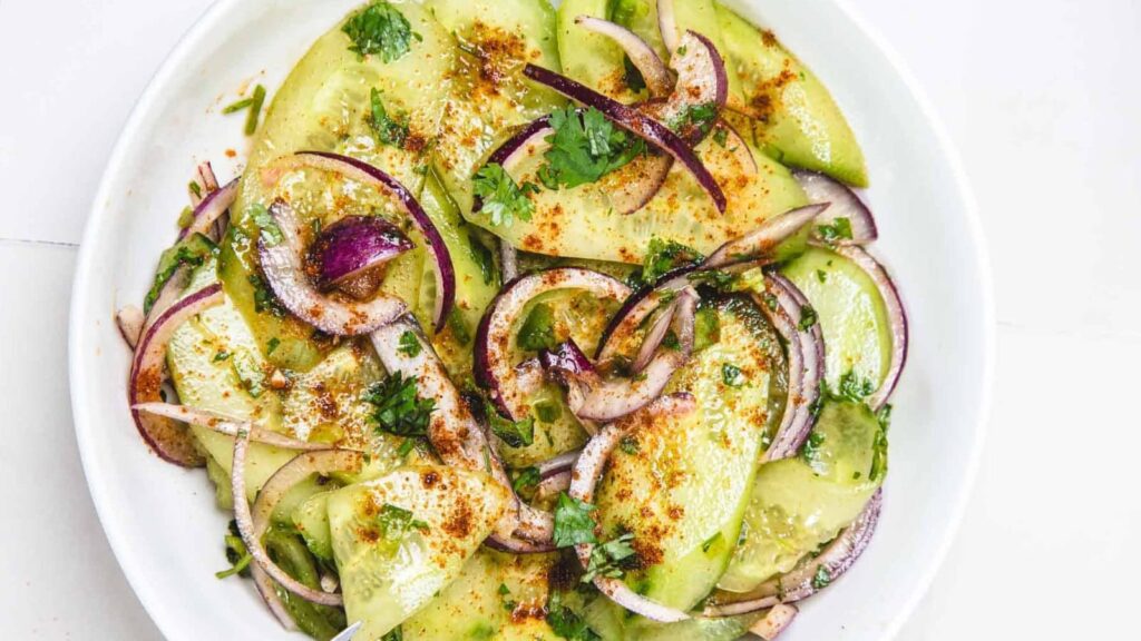 Sliced cucumber and red onion salad topped with herbs and spices on a white plate.