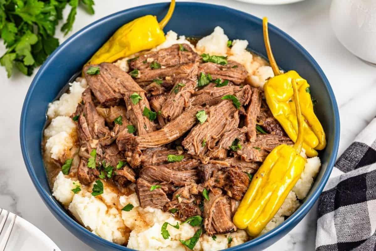 A blue bowl filled with shredded pot roast on mashed potatoes, garnished with parsley and whole yellow peppers.