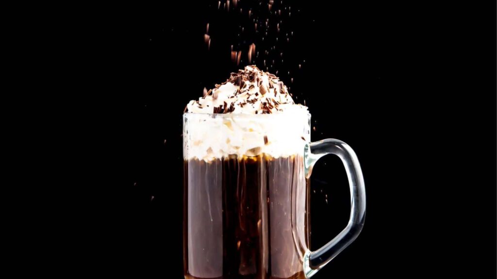 A glass mug of hot chocolate topped with whipped cream and chocolate shavings, with more shavings sprinkling down, set against a dark background.