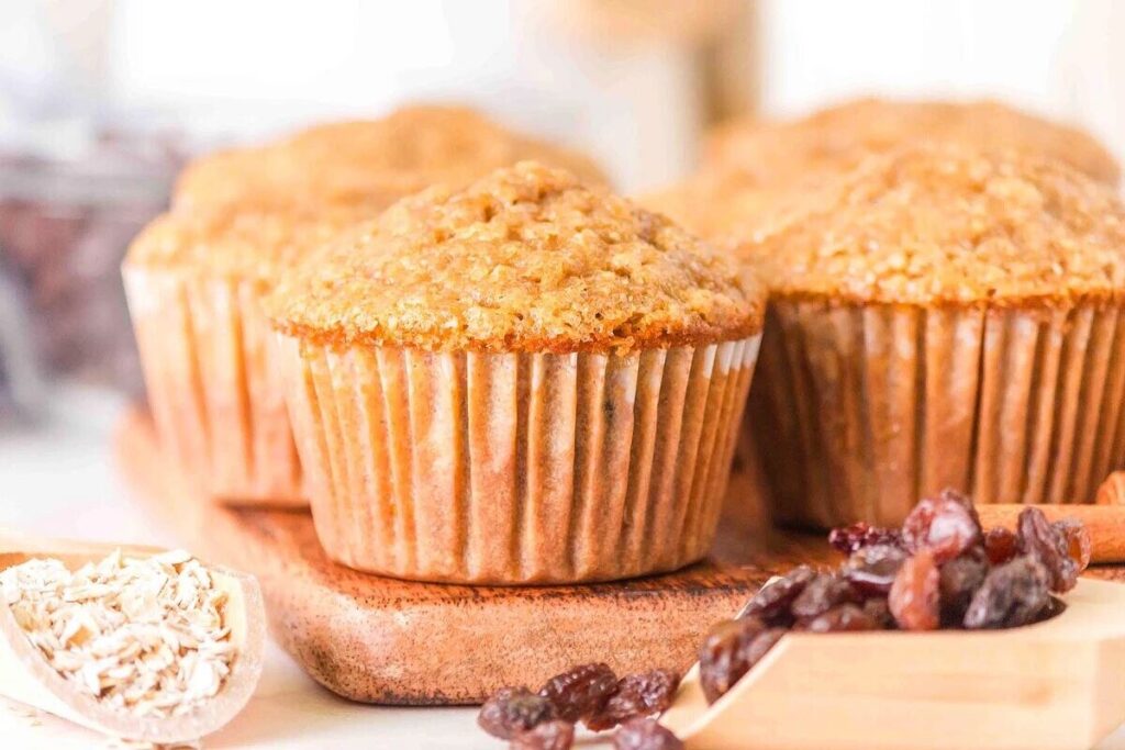 Four freshly baked oatmeal raisin muffins are arranged on a wooden board, surrounded by a spoonful of oats and scattered raisins.