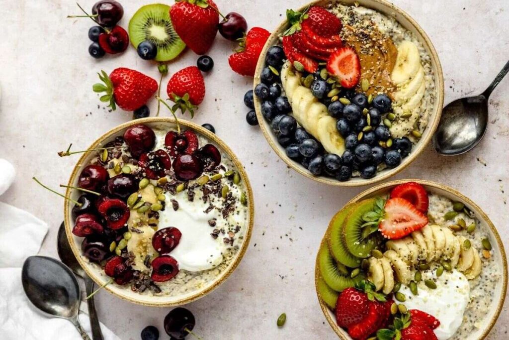 hree bowls of healthy oatmeal breakfast are topped with fresh fruit, seeds, and nuts, surrounded by spoons and assorted fruit.