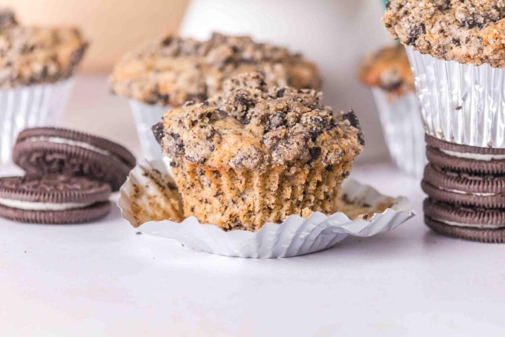 A close-up of a crumbled Oreo muffin, partially unwrapped from its foil liner, with two stacks of chocolate sandwich cookies in the background. Other muffins are visible but out of focus.