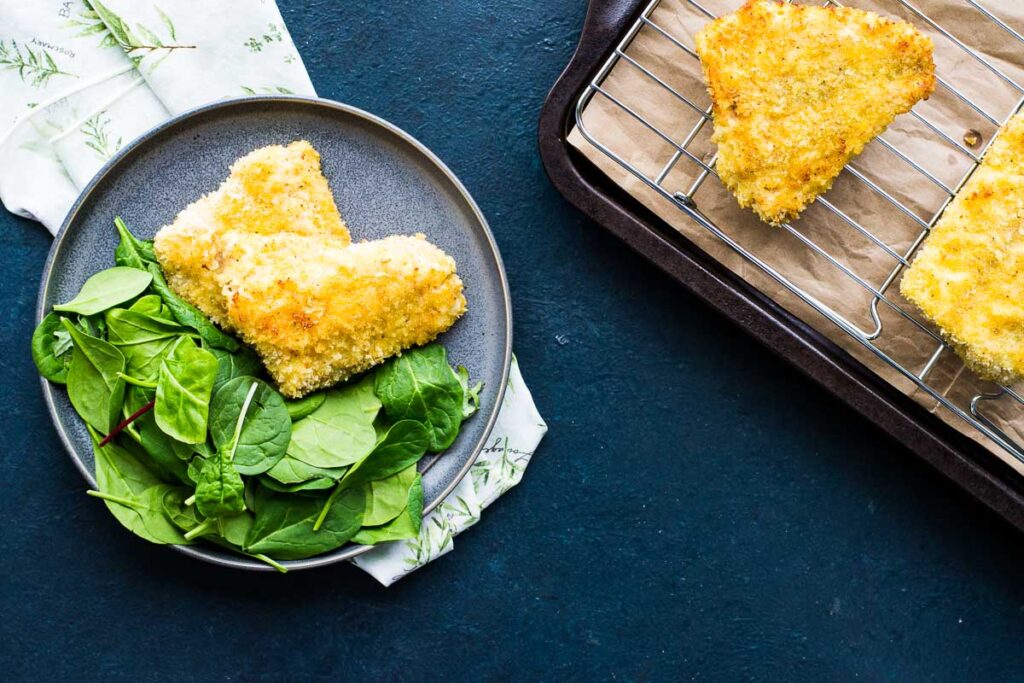 A panko crusted rockfish on a plate with spinach next to a serving platter with more crispy fried fish.
