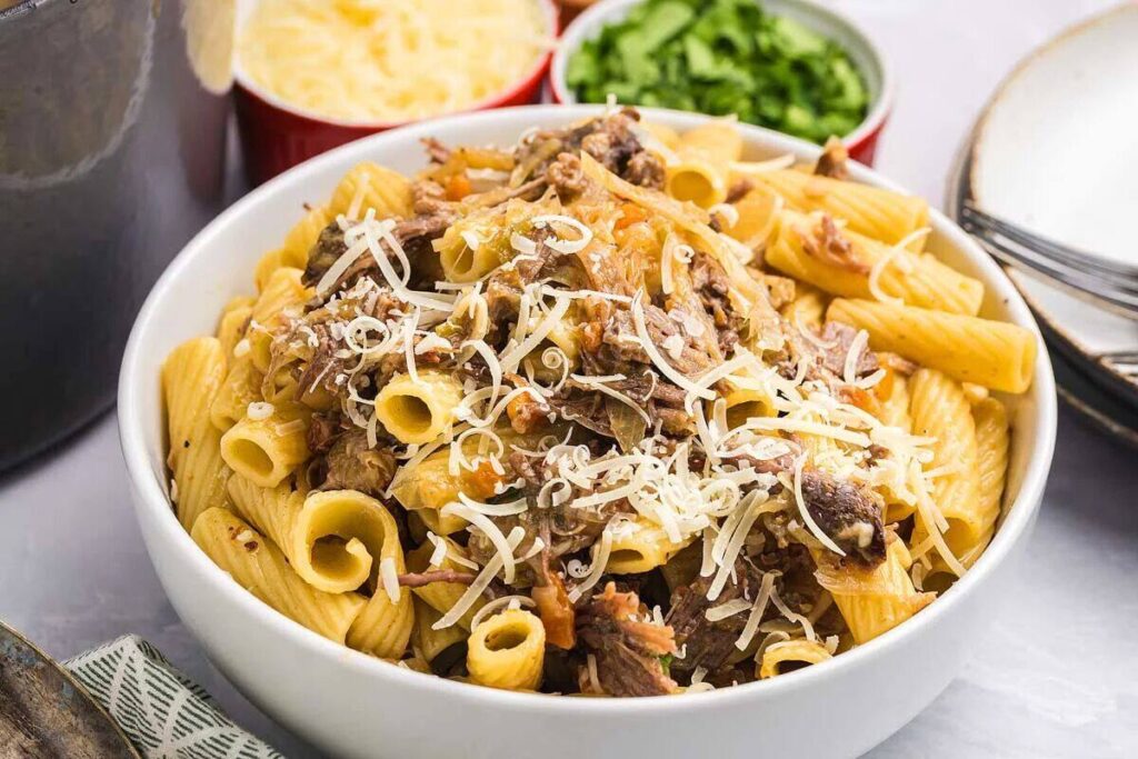 Rigatoni pasta alla Genovese topped with shredded meat and grated cheese, with bowls of cheese and herbs in the background.