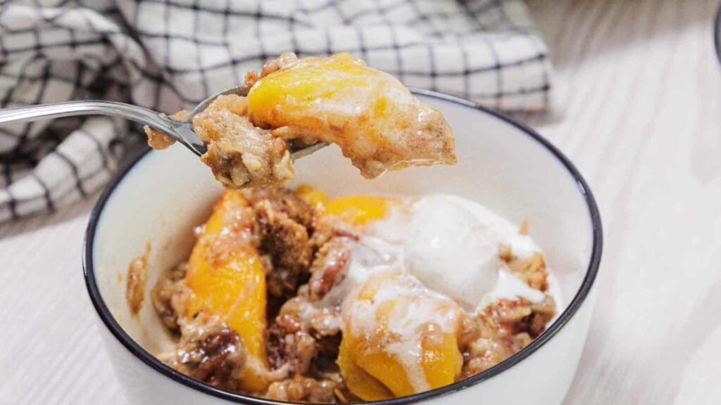 A bowl of peach cobbler topped with melted ice cream sits on a table; a spoonful of cobbler with peaches and crumbly topping is held above the bowl. A checkered cloth is in the background.