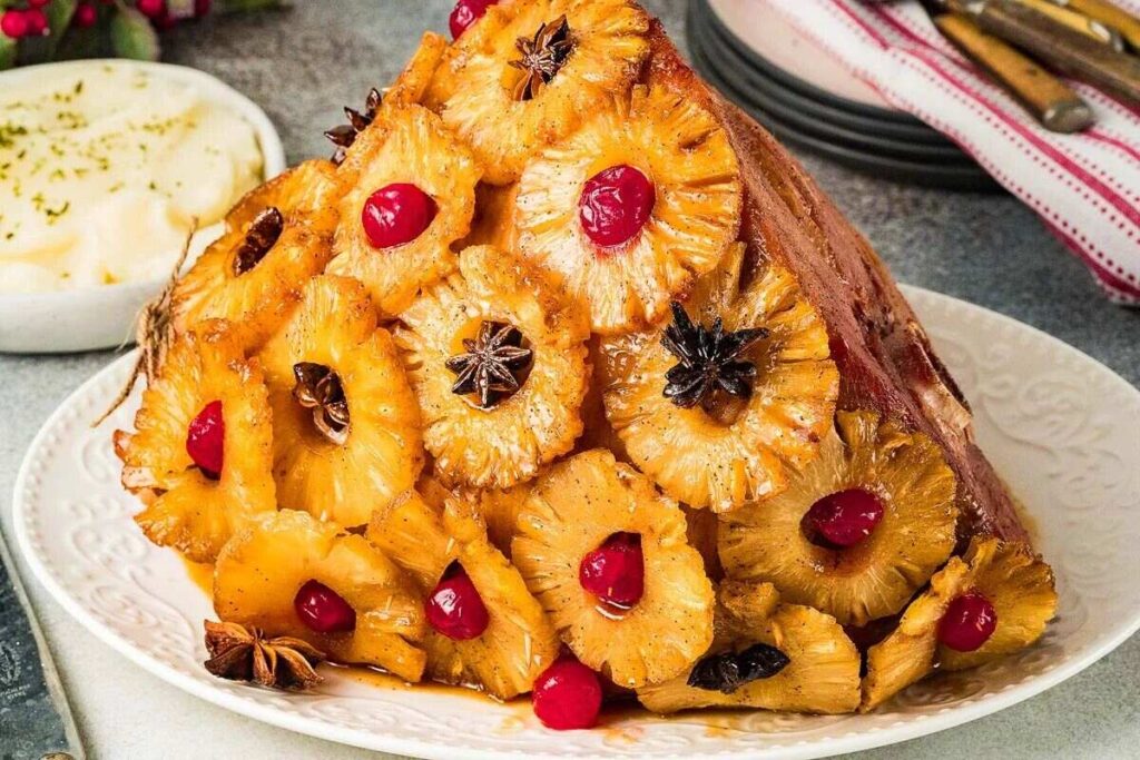 A brown sugar pineapple glazed ham garnished with pineapple slices, cherries, and star anise on a white plate, with a bowl of mashed potatoes and utensils in the background.
