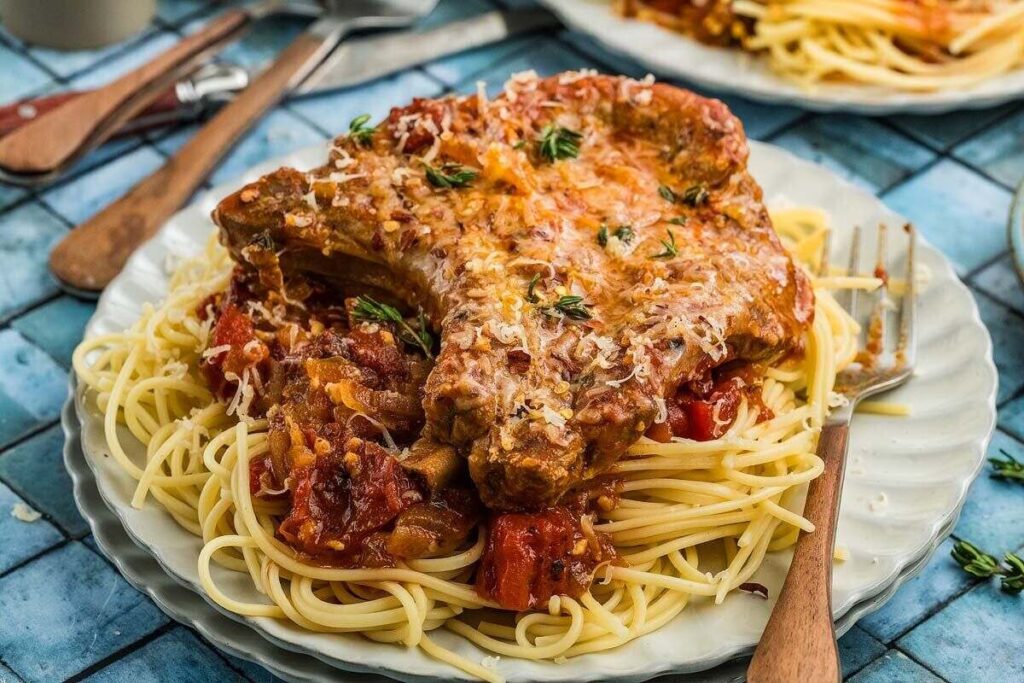 Pork chops Italiano with savory tomato sauce served over spaghetti on a white plate, with a fork and knife on the side.