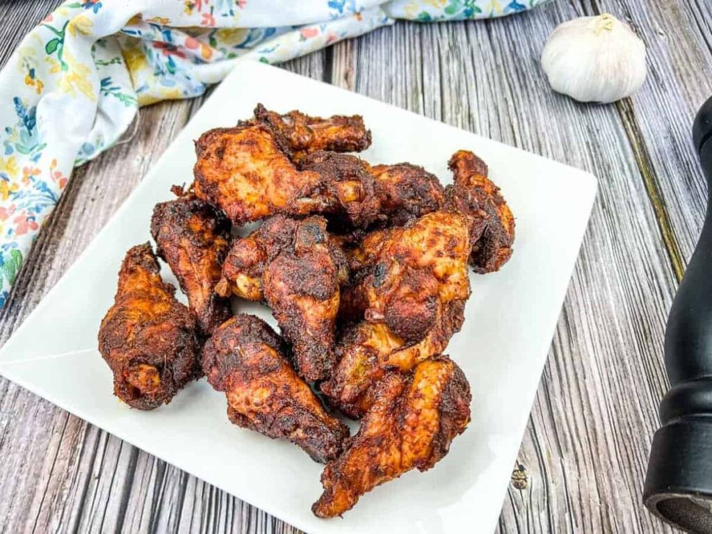 A white square plate with several pieces of seasoned, cooked chicken wings sits on a wooden table, next to a garlic bulb, a pepper grinder, and a floral cloth.