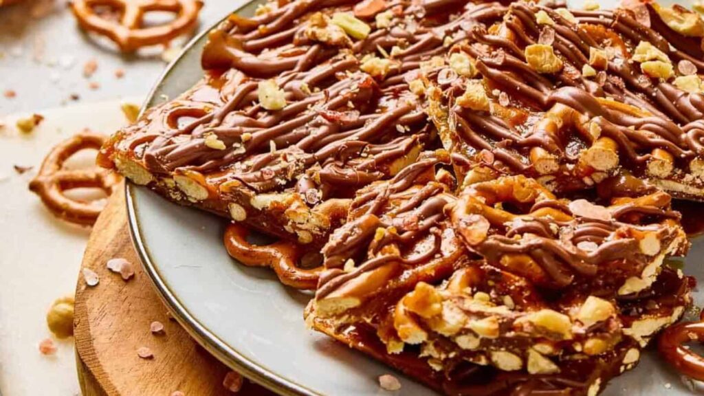 Chocolate-drizzled pretzel toffee pieces on a gray plate, topped with chopped pretzels and nuts.