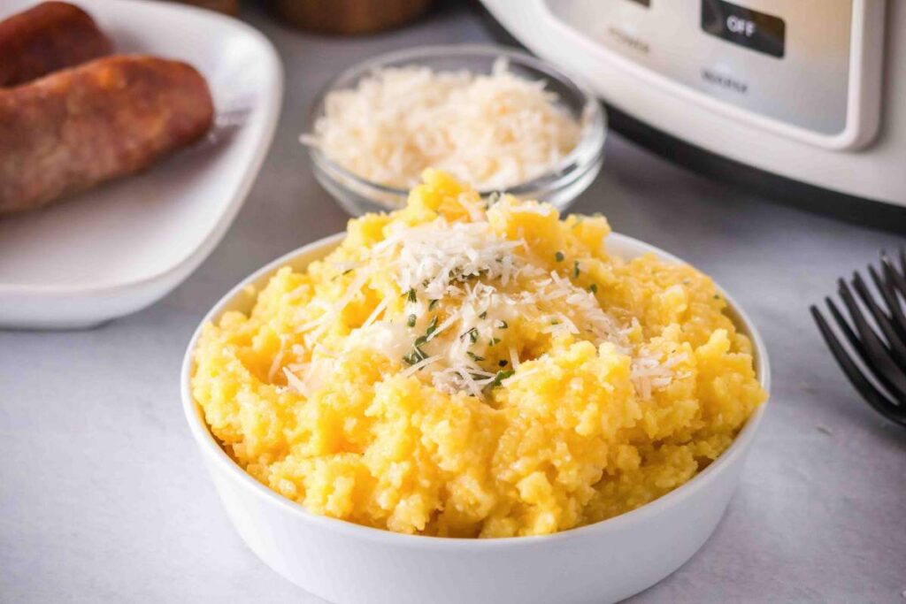 A bowl of creamy polenta topped with grated parmesan cheese and herbs, served next to sausages and a slow cooker in the background.