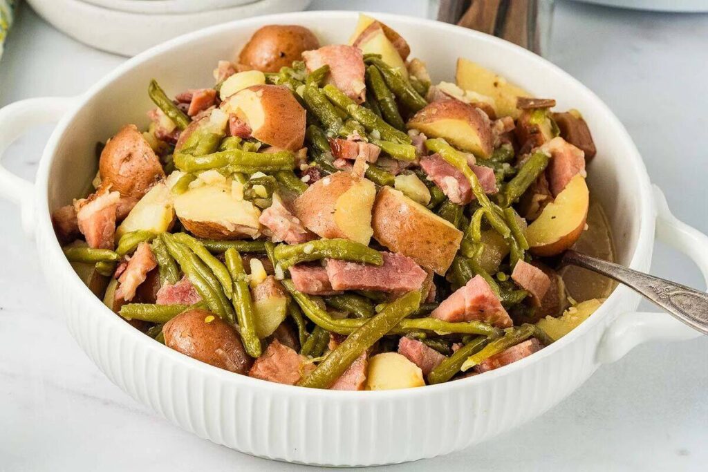 A white dish filled with a mixed serving of slow cooker ham, green beans, and red potatoes. A spoon is placed inside the dish.