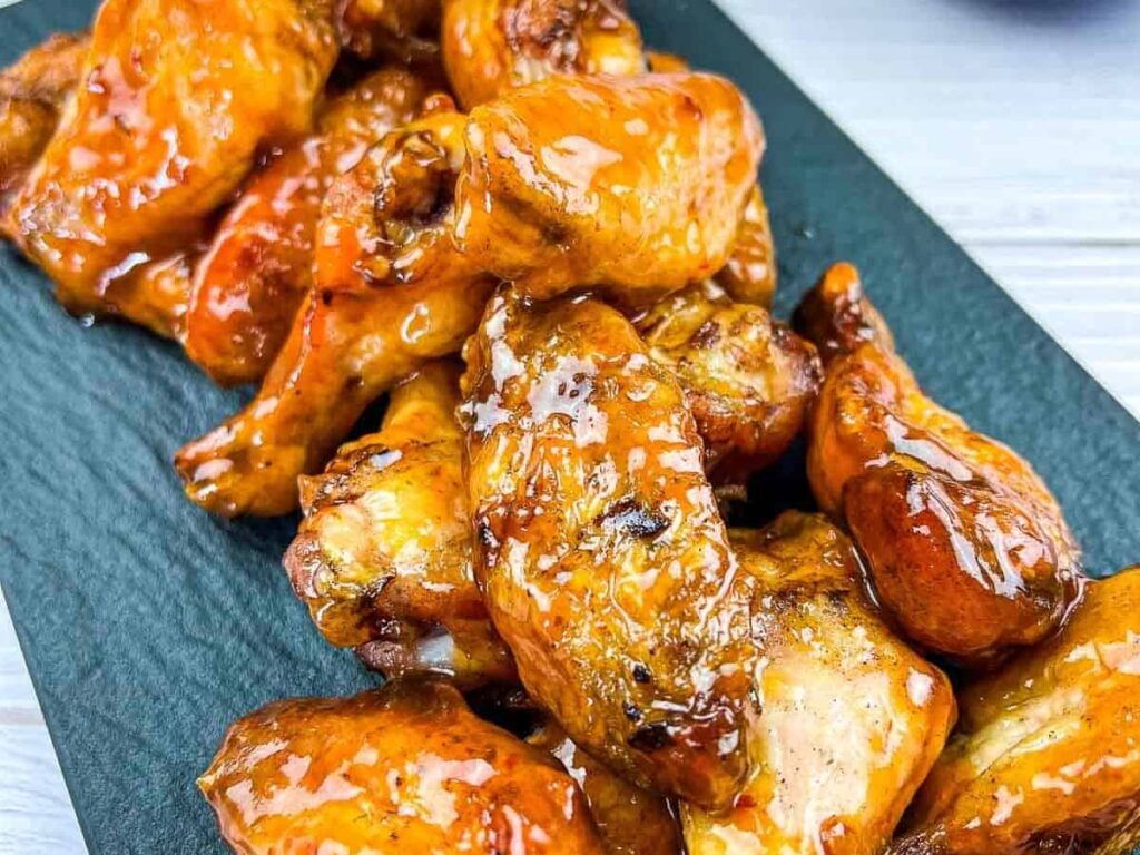 Close-up of a pile of glazed peach-chipotle chicken wings with crispy skin, glistening in a sweet and savory sauce, on a dark rectangular serving platter.