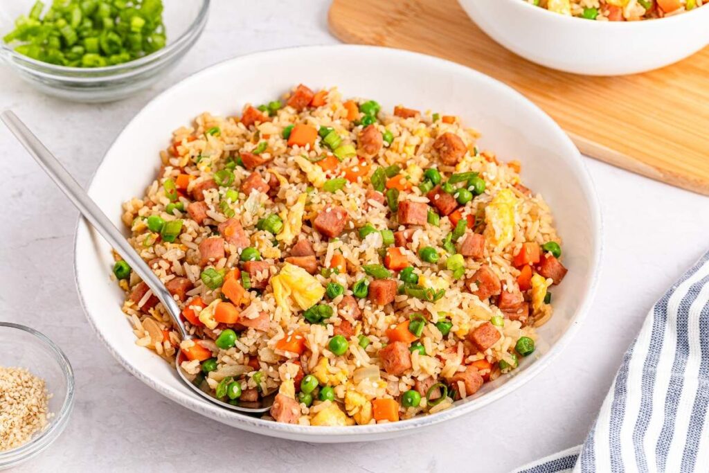 A white bowl filled with spam fried rice containing diced ham, peas, carrots, and scrambled eggs is on a table. 