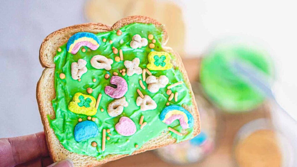 Two slices of bread with green icing and colorful cereal pieces on a wooden cutting board. A textured yellow cloth is partially visible on the table, alongside a bowl of cereal.