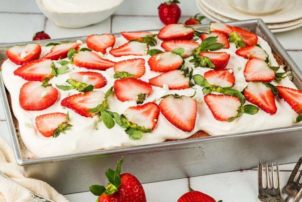 In a metal pan, this delightful strawberry tres leches cake is topped with whipped cream and sliced strawberries. Beside it, a few whole strawberries and a fork await to enhance the indulgence.