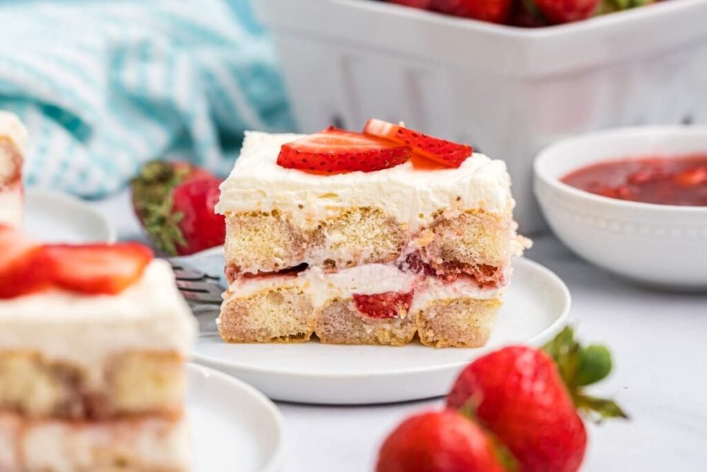 A luscious slice of strawberry tiramisu graces a white plate, adorned with fresh sliced strawberries. In the background, extra strawberries and a bowl of vibrant strawberry sauce complete this delightful scene.