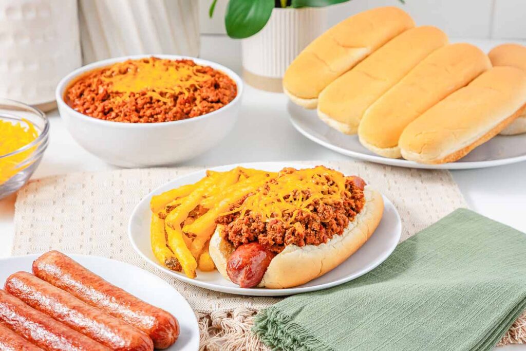 Chili cheese hot dog with French fries on a white plate, set on a beige placemat next to a green napkin.