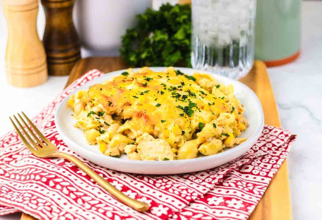 A plate of cheesy pasta casserole garnished with chopped parsley, on a red-patterned napkin with a gold fork.