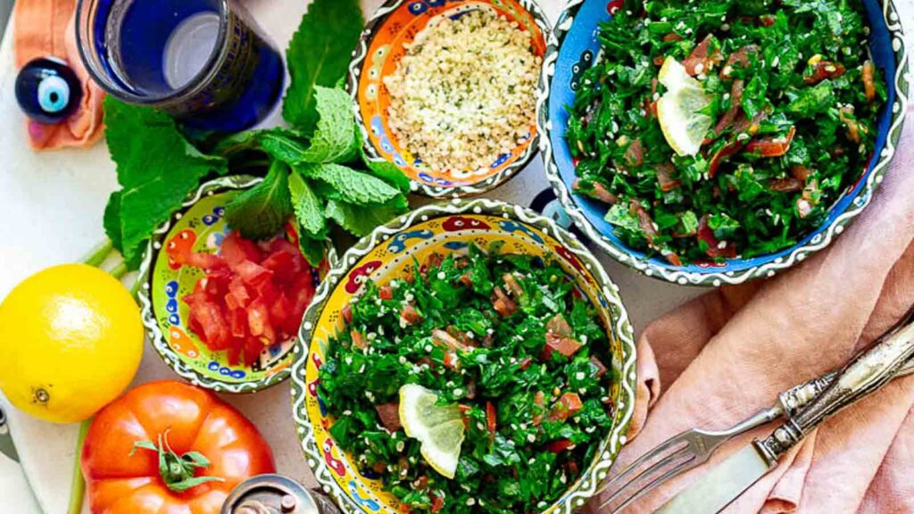 Overhead shot of a table setting featuring multiple bowls of tabbouleh salad, lemon, tomato, and mint leaves, with decorative bowls, a blue glass, and cutlery.