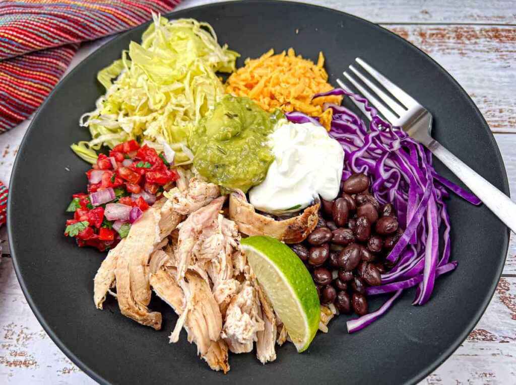 A black ceramic bowl filled with a deconstructed chicken burrito bowl, featuring shredded chicken, pico de gallo, shredded lettuce, shredded cheddar cheese, guacamole, sour cream, black beans, shredded red cabbage, and a lime wedge, with a fork resting on the plate.