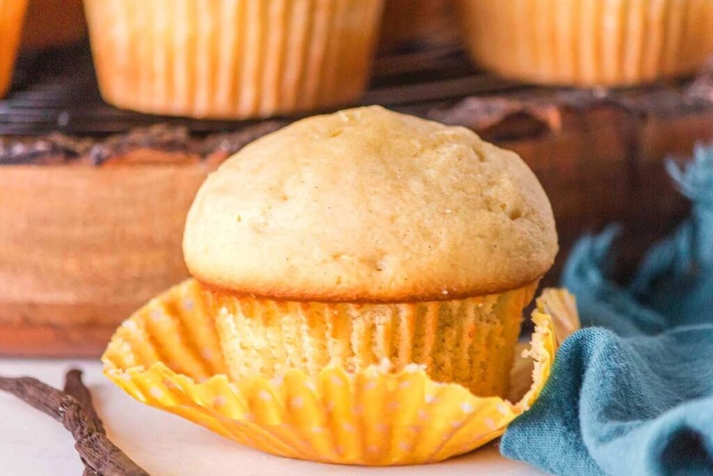 A single vanilla muffin partially unwrapped from a yellow polka dot paper liner, with more vanilla muffins in the background on a wooden surface.